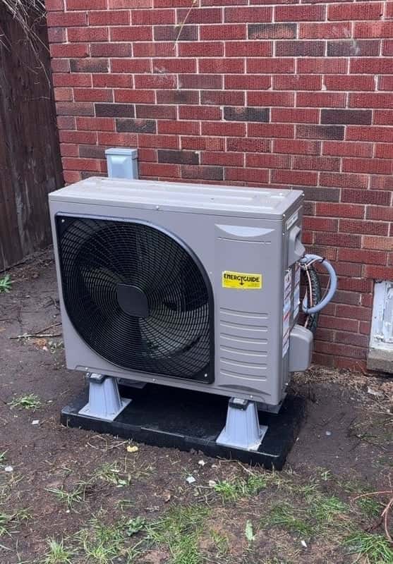 Outdoor heat pump unit installed on a raised pad against a brick wall